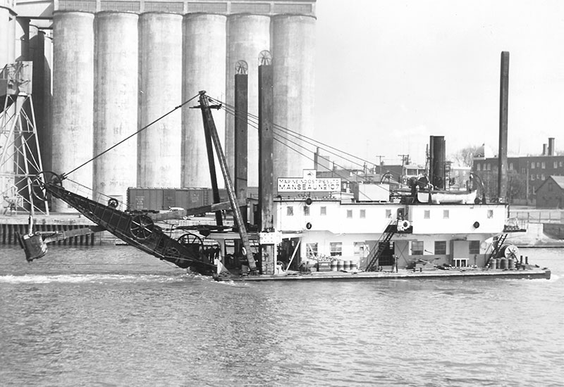 On voit la drague <em>Manseau 107</em> sur la rivière Richelieu près des silos à grains