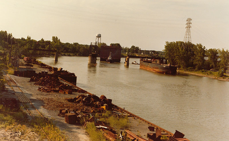La cale sèche est remorquée sur la rivière Richelieu entre les piliers du pont de la voie ferrée.