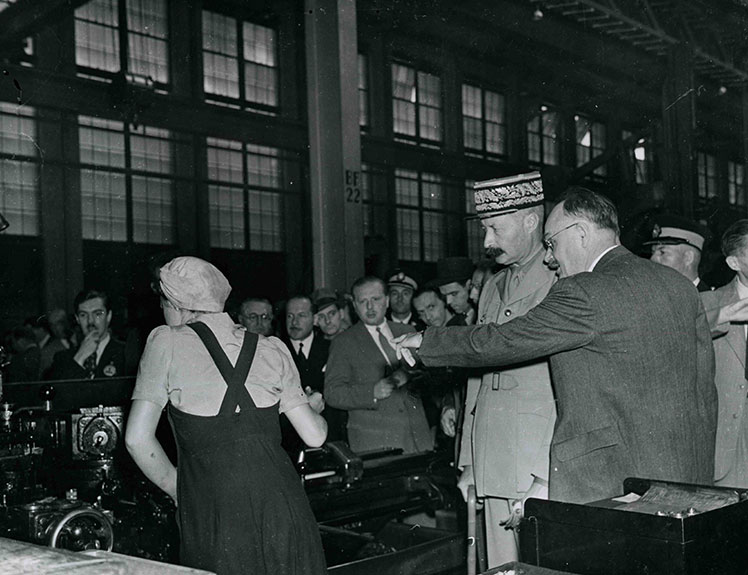 Un homme portant un képi regardant une ouvrière à l'œuvre.