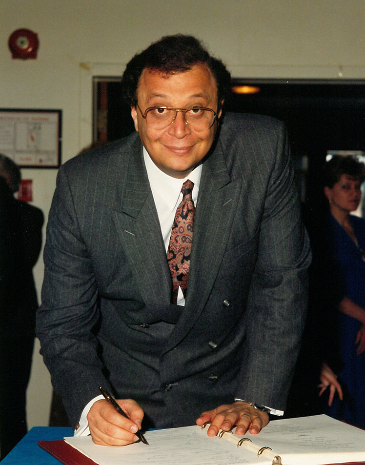 Un homme vêtu d'un habit et portant des lunettes signant un livre.